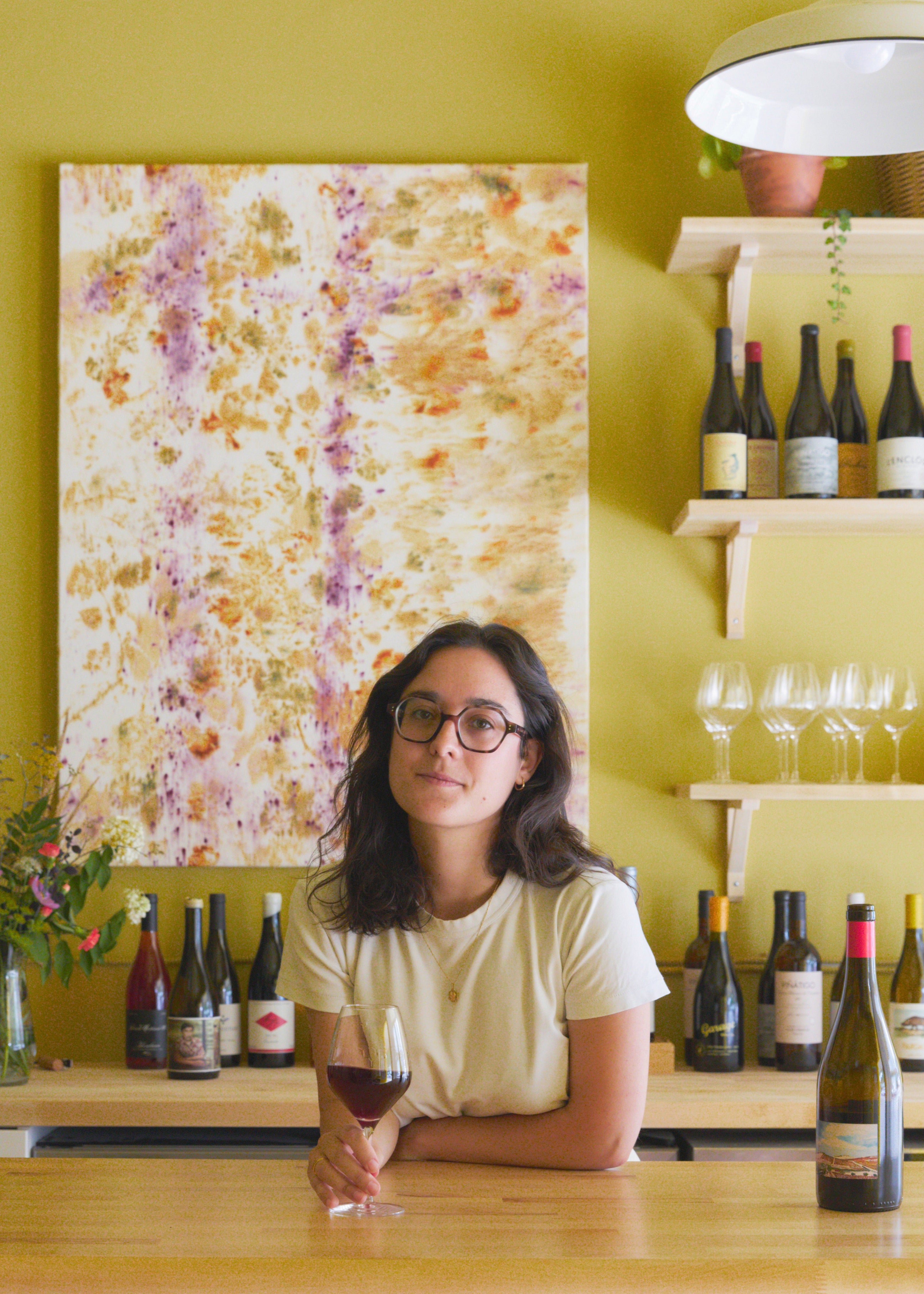 Celia holding a glass of wine in at Canary Counter with bottles and Nathalie Leturcq artwork on the wall.