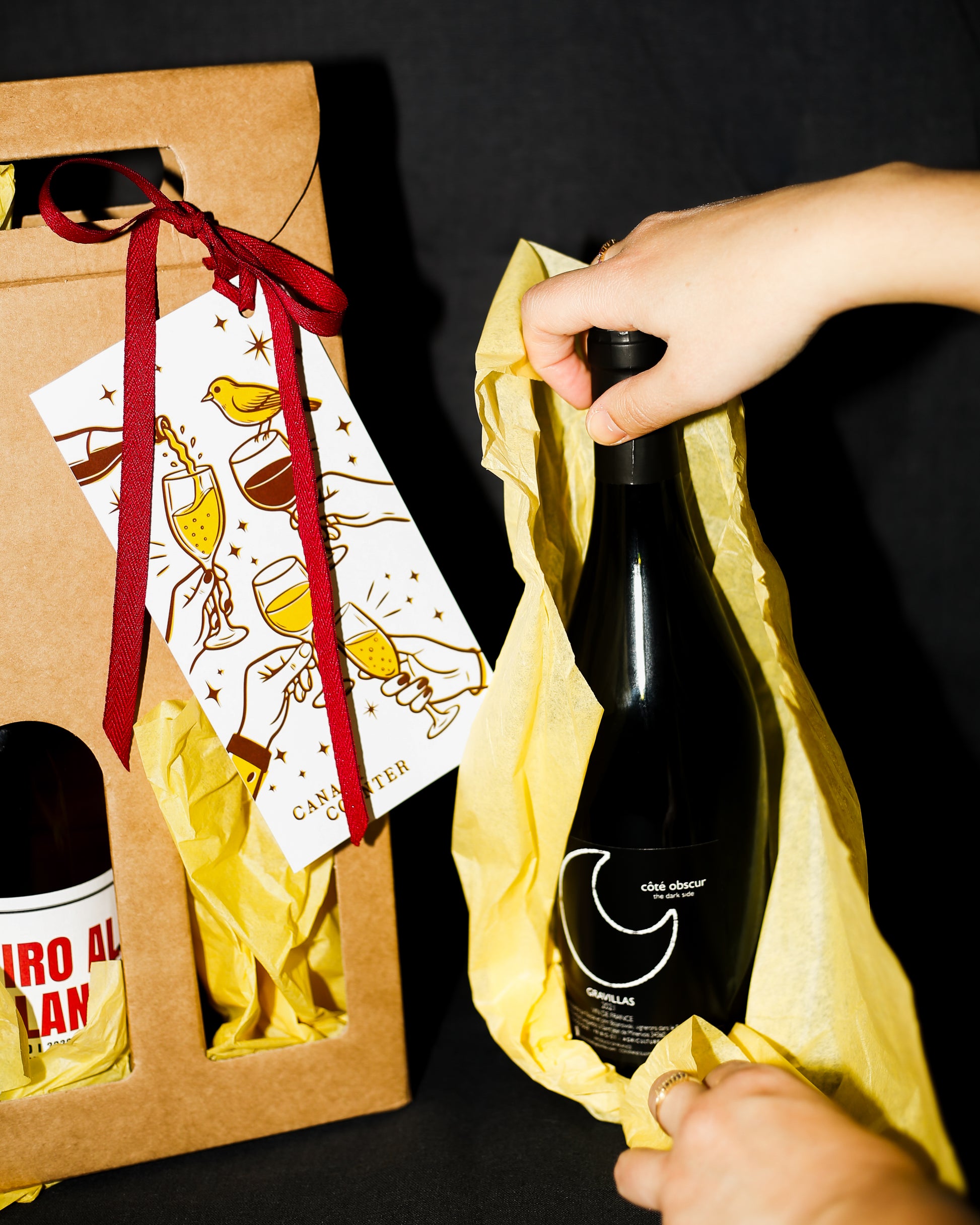 Person holding a bottle of wine with a gift box featuring a red ribbon and card in the background.
